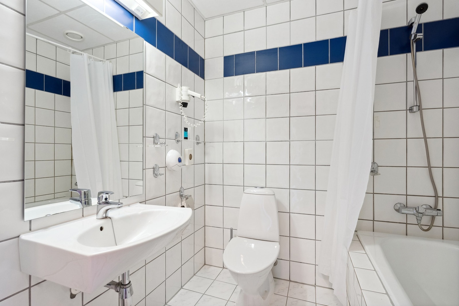 Bad med flisbelagte overflater, med varmekabler i gulvet. Det er montert vegghengt porselenservant, gulvstående toalett, vegghengt speil og badekar med dusjgarnityr på vegg. Galleribilde