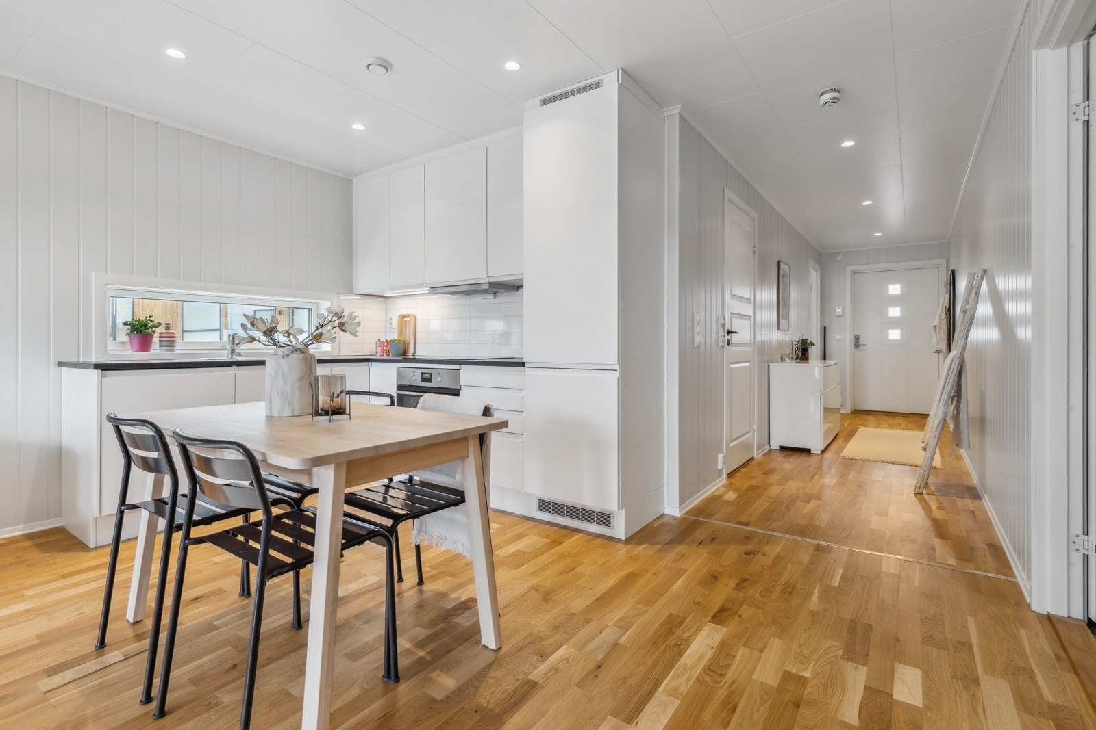 Stilrent og moderne kjøkken fra Aubo i åpen løsning mot stuen. Innredning med hvite slette fronter med foring opp til tak og laminert benkeplate i mørk utførelse med nedfelt oppvaskkum i rustfritt stål. Galleribilde