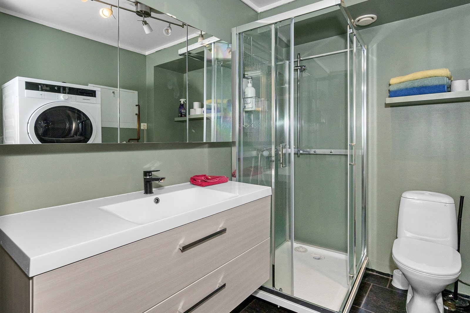 Et stort og godt baderom/vaskerom med dusjkabinett, innredning med nedfelt vask, underskap, toalett, speil, vaskemaskin og tørketrommel Galleribilde