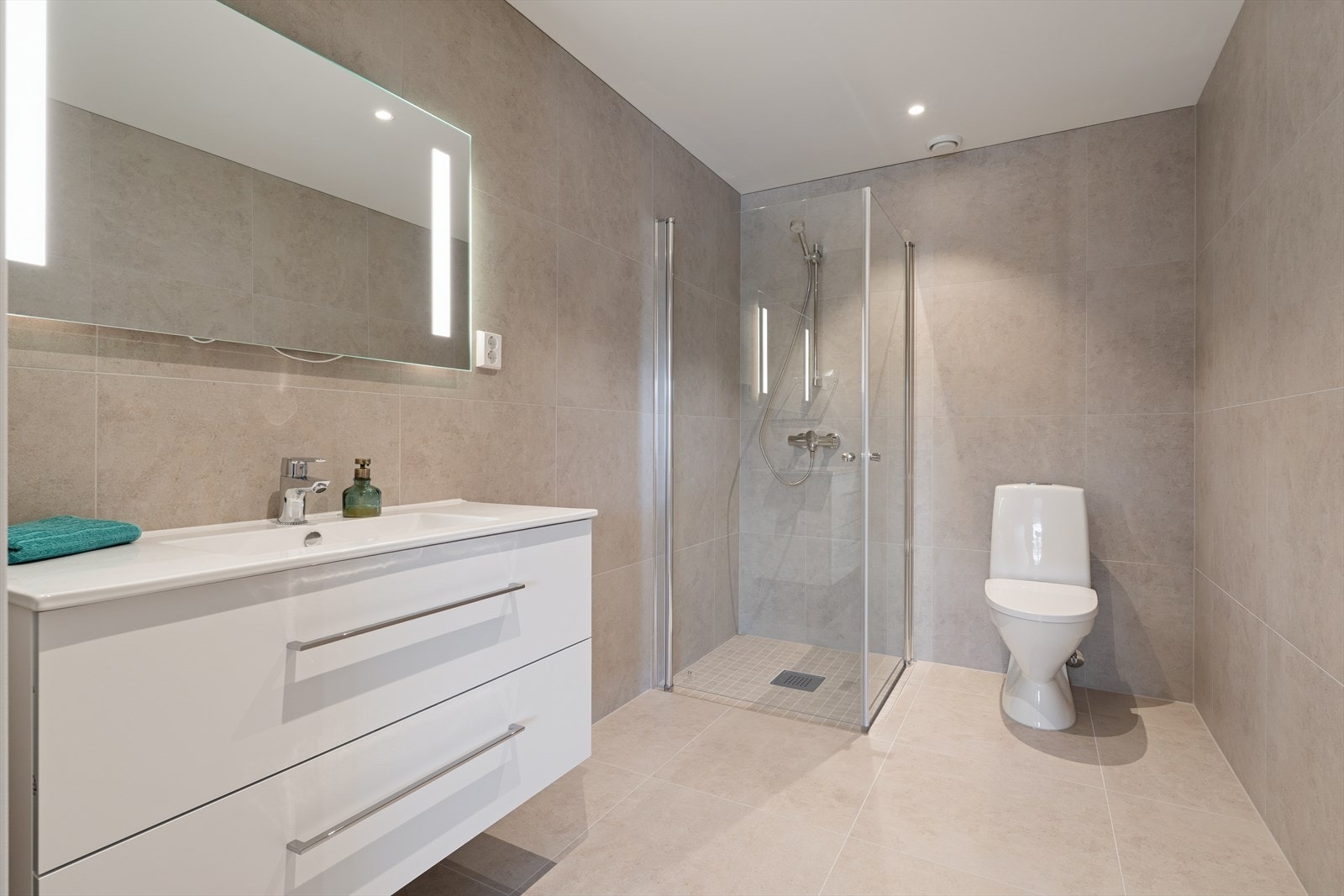 Badet har fliser på gulv og vegger, med malte gipsplater og innfelte spotter i himlingen. Det er vannbåren gulvvarme for økt komfort. Galleribilde