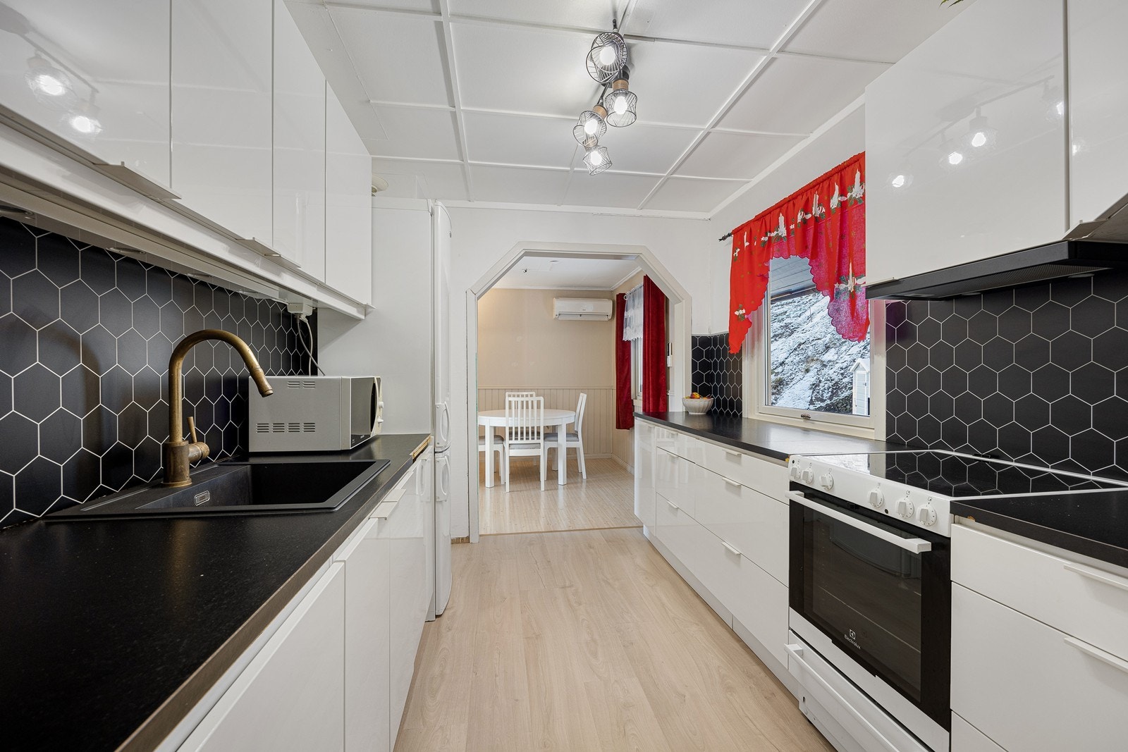 Over kokesonen er en ventilator etablert, og over benken har kjøkkenet belysning, stikkontakter og sorte kitchenboards med heksagonmønster. Gulvet har 1-stavs laminat, og to parallelle seksjoner gir god skap- og benkeplass. Galleribilde