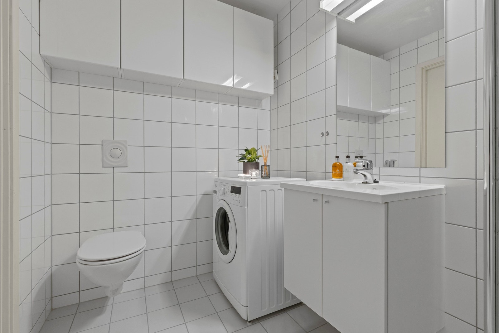 Badet er utstyrt med veggmontert toalett, dusjhjørne vegg/dør i herdet glass, servant, skap, speil og belysning og opplegg for vaskemaskin. Galleribilde