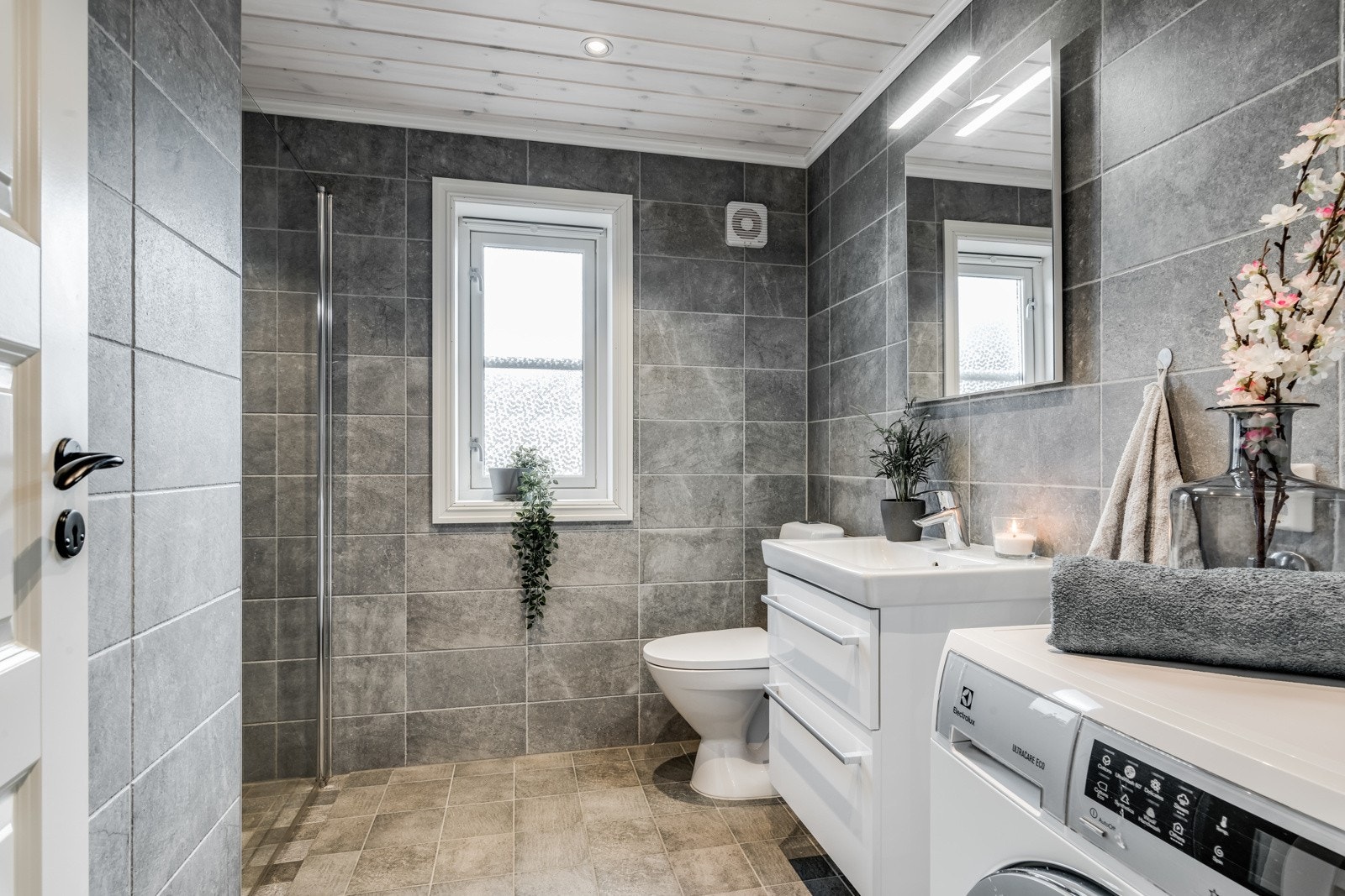 Flott baderom med moderne fliser, helservant på skuffeinnredning, klosett, dusjnisje og opplegg for vaskemaskin. Galleribilde