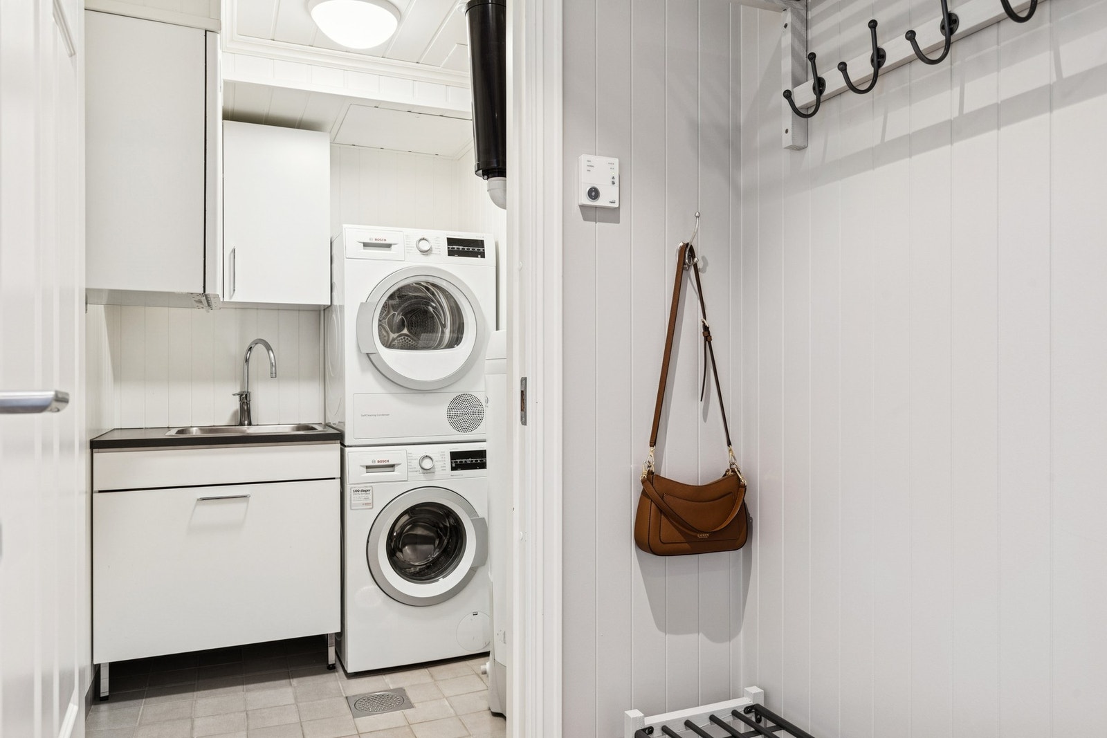 Praktisk vaskerom med flislagt gulv og gulvvarme. Rommet er innredet med benkeskap og benkeplate, utslagsvask og opplegg for vaskemaskin og tørketrommel. Galleribilde