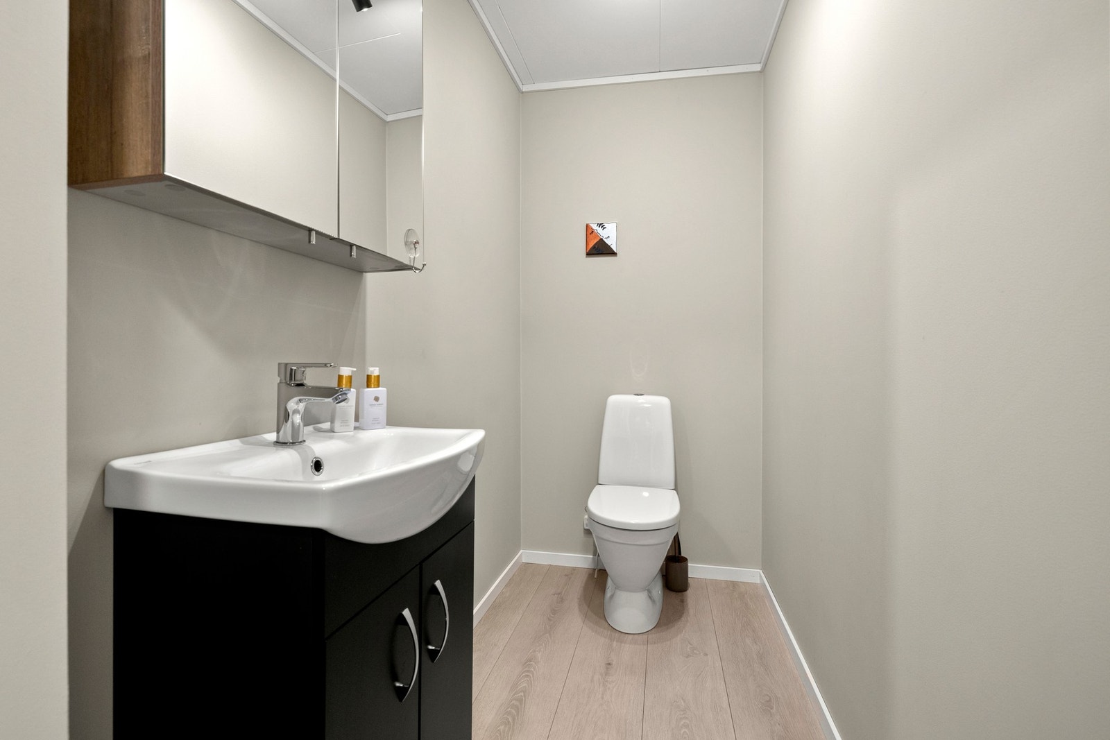 Lyst toalettrom med sort servantskap, speilskap og gulvmontert toalett. Parkett på gulv og malte vegger i samme lyse tone som resten av etasjen. Galleribilde