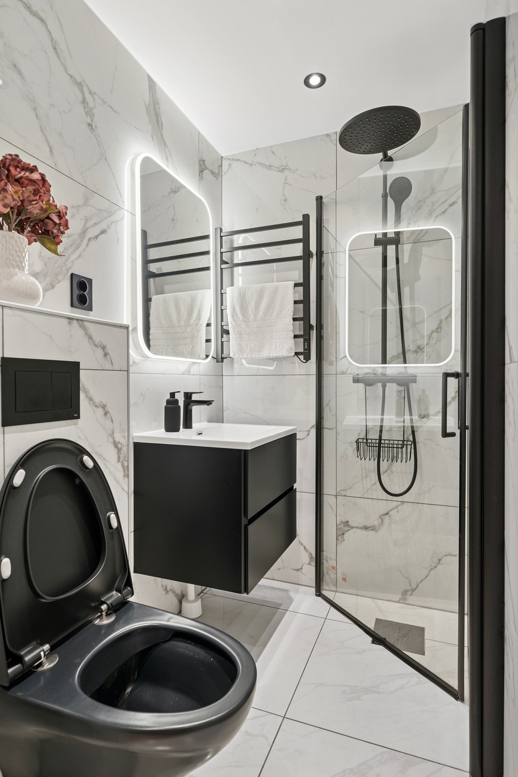 Sort vegghengt toalett med innebygget sisterne. Servantinnredning med sorte glatte fronter, hvit heldekkende servant, sort ettgreps servantarmatur, speil med led belysning. Sort elektrisk håndkletørker. Dusjhjørne rett på gulv med innfellbare glassdører. Galleribilde