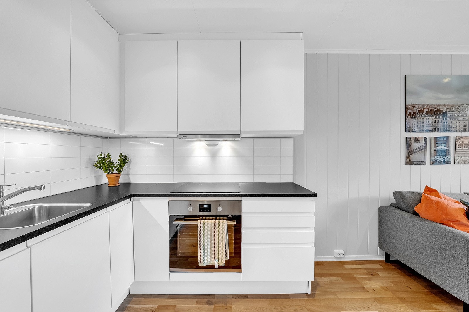 Kjøkkenet fra Aubo er moderne og praktisk utformet med hvite fronter og en mørk benkeplate som skaper en elegant kontrast. Det er belysning under overskap og plate på vegg over benkeskap. Galleribilde