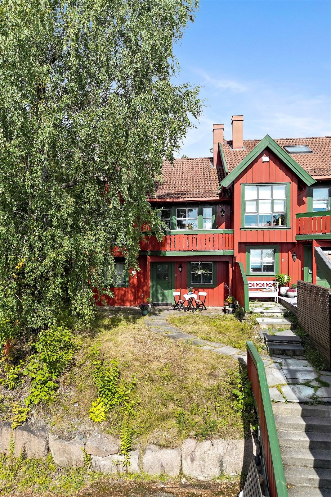 Boligen har adkomst fra en solrik og hyggelig uteplass med skifer, gressplen og frodige trær Galleribilde