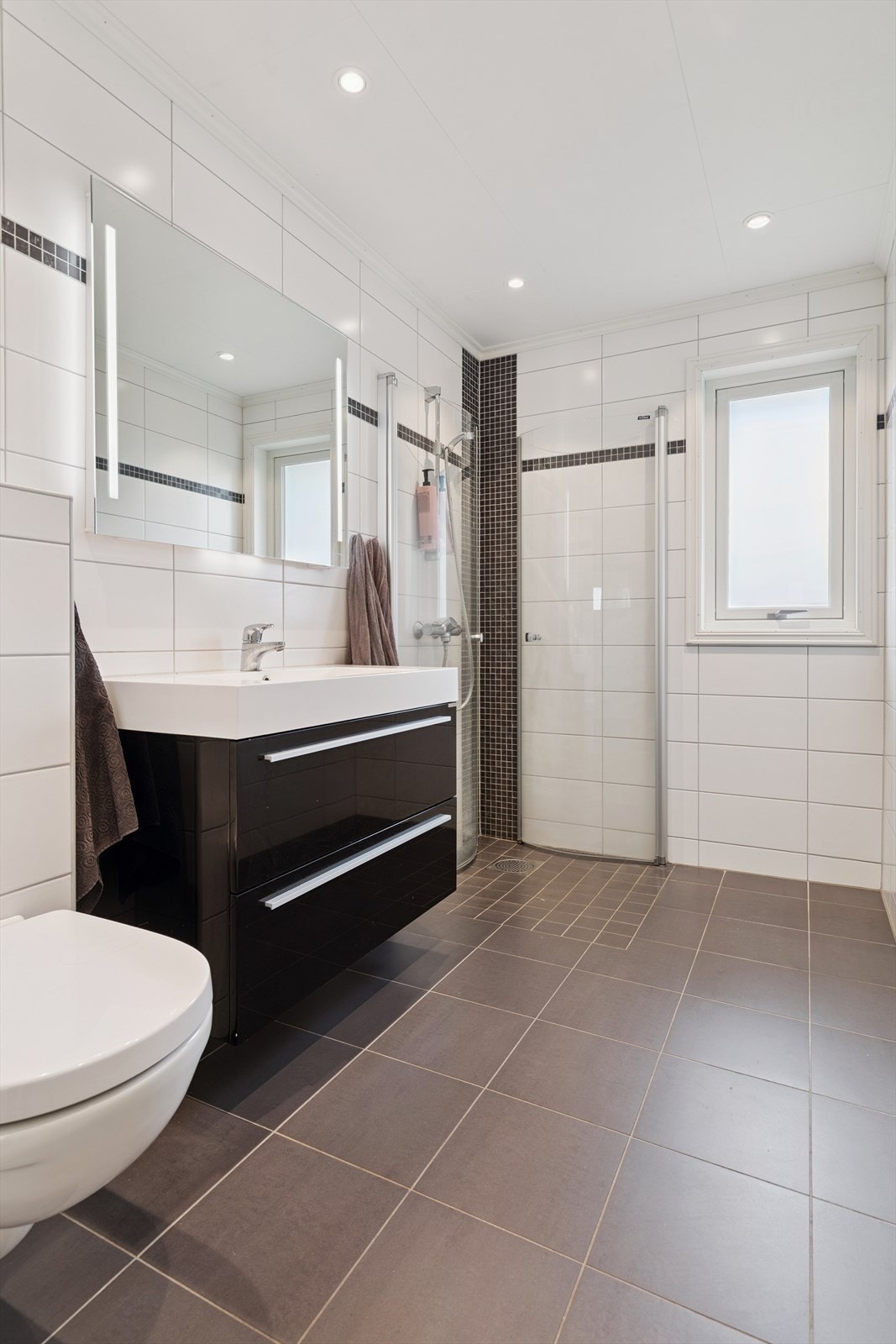 Bad i hovedetasjen med flislagt gulv og vegger, dusjhjørne og servant med skuffeseksjon. Galleribilde