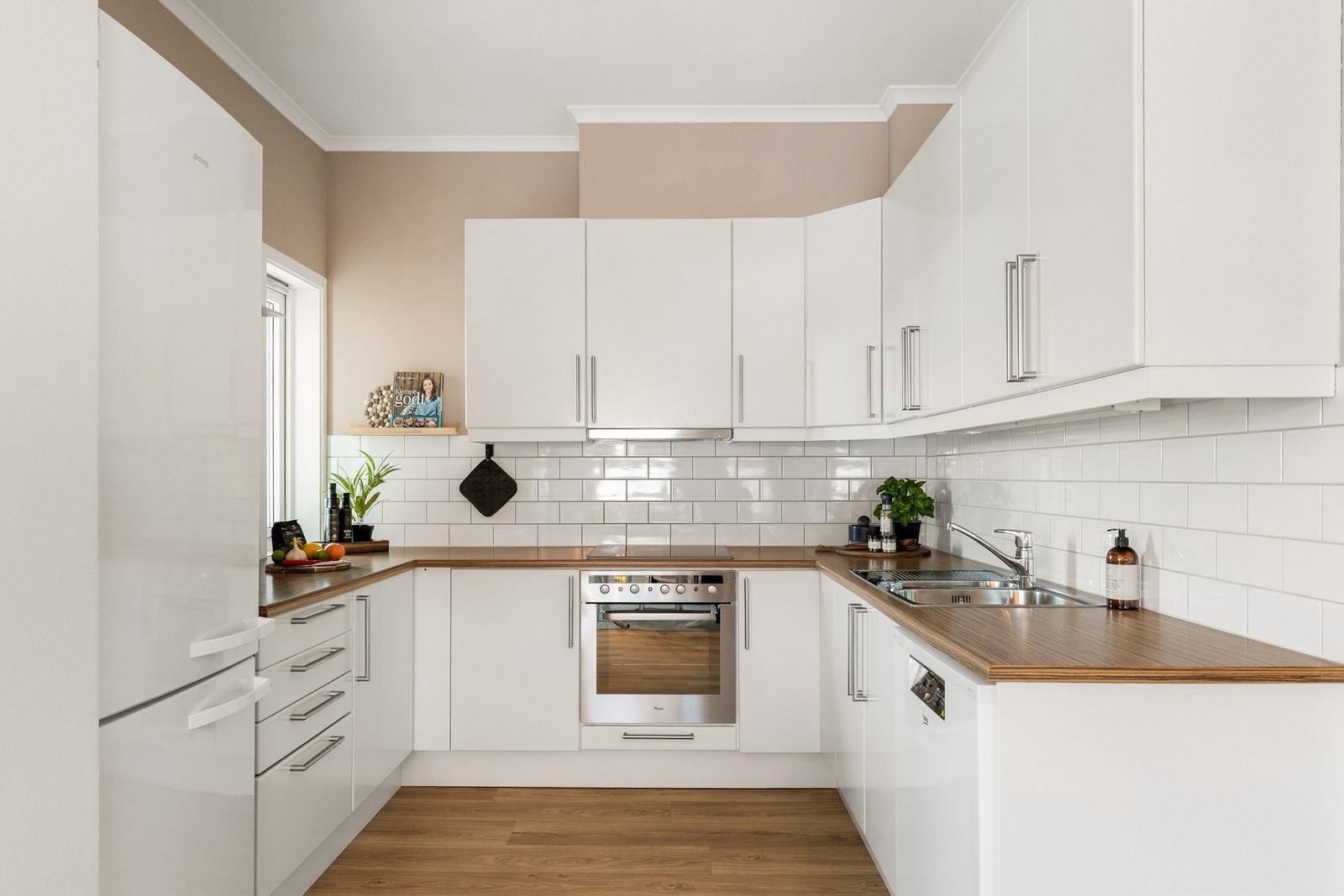 Kjøkkeninnredning fra Sigdal. Det er montert hvite, slette fronter og benkeplater i laminat. Det er montert flis mellom overskap og benkeplate. Keramisk koketopp, integrert stekeovn og oppvaskmaskin. Det er også nisje for frittstående kjøleskap. Galleribilde