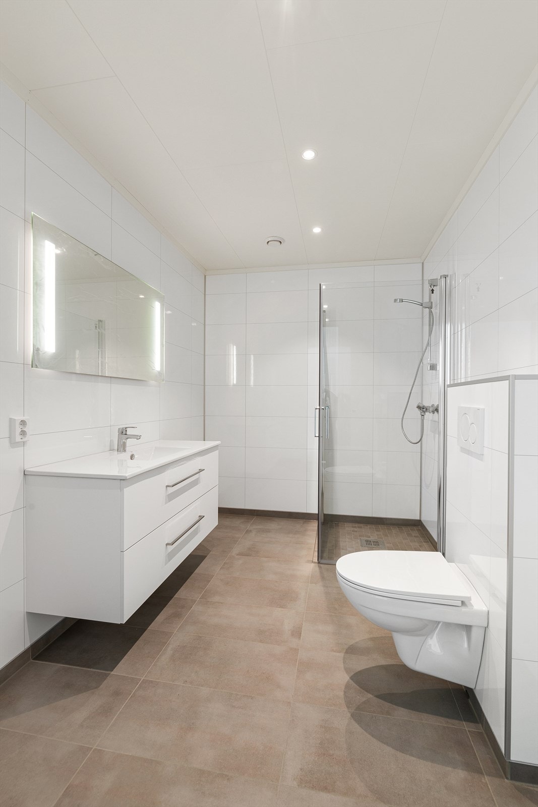 Hvit baderomsinnredning med heldekkende nedfelt servant og rom for oppbevaring i skuffeseksjon, speil med belysning, dusjhjørne og vegghengt toalett. Galleribilde