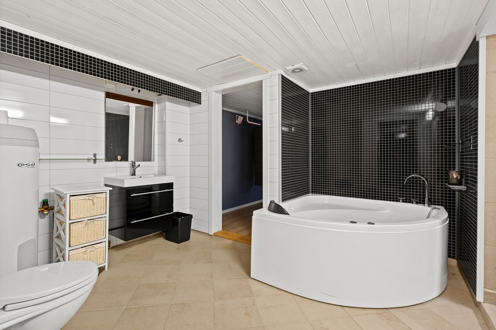 Badet har hvite og svarte fliser, et hjørnebadekar, servant med speil og toalett. Galleribilde