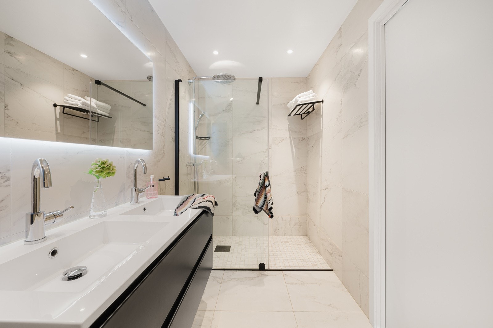 Badet er innredet vegghengt toalett, dusjnisje med glass skyvedør, dobbel servant med møblement og overhengende speil. Galleribilde