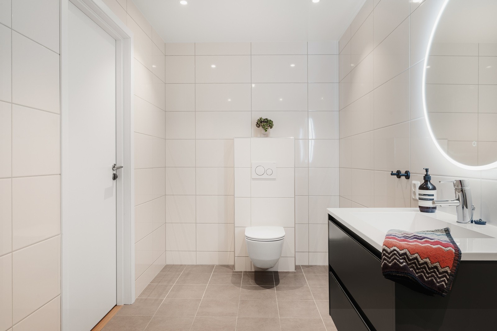 Badet er innredet med vegghengt toalett, dusj med glass foldedører, servant med møblement, overhengende speil og belysning. Galleribilde