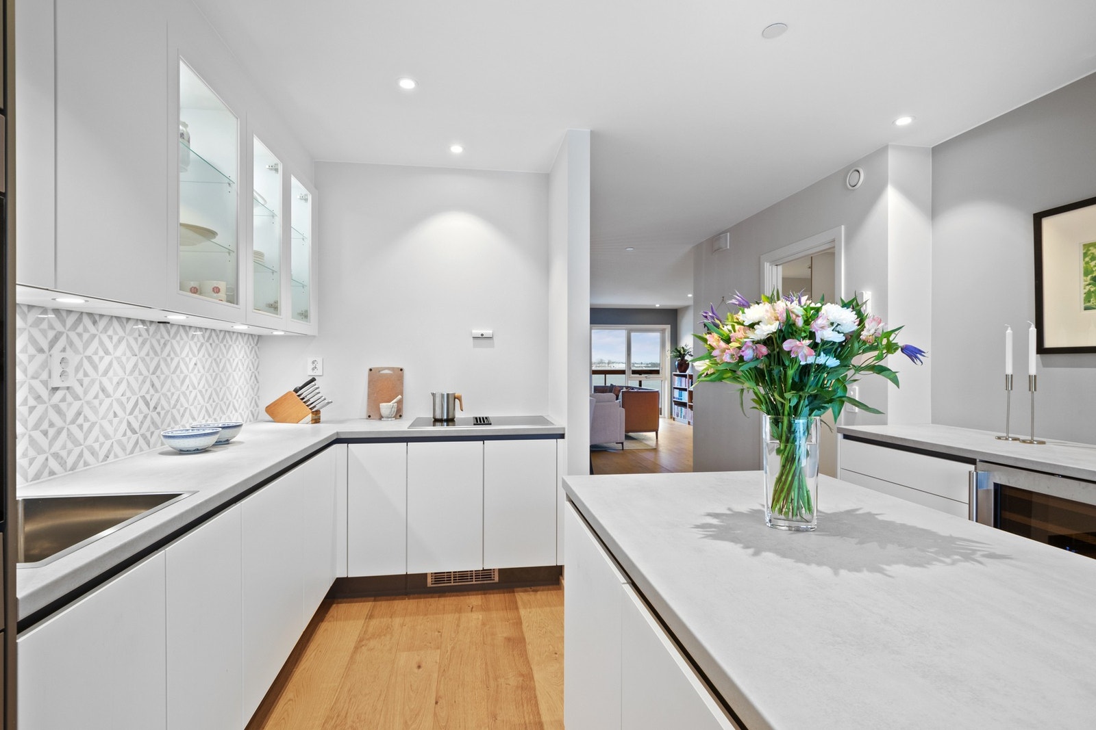 Innredningen er stilren og moderne, med hvite, glatte fronter, vitrineskap, mørkegrå høyskap, nedfelt oppvaskkum og lysegrå benkeplate av laminat. Galleribilde