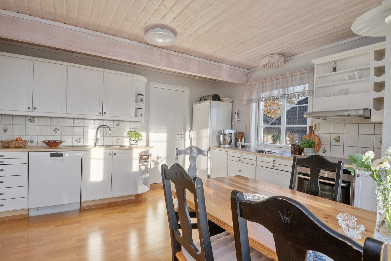 Over benken har kjøkkenet hvite fliser med noe dekor, og over kokesonen er en ventilator etablert med avtrekk ut. Galleribilde