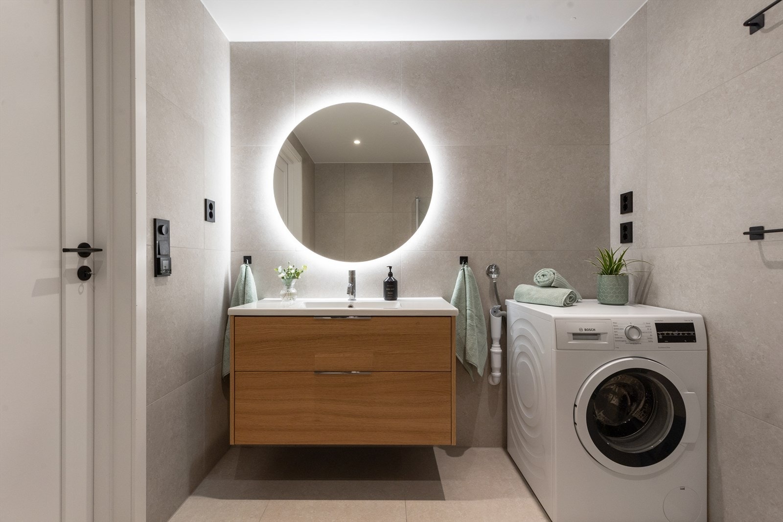 Delikat flislagt bad med servantinnredning, bakbelyst speil og opplegg for vaskemaskin og tørketrommel. Galleribilde