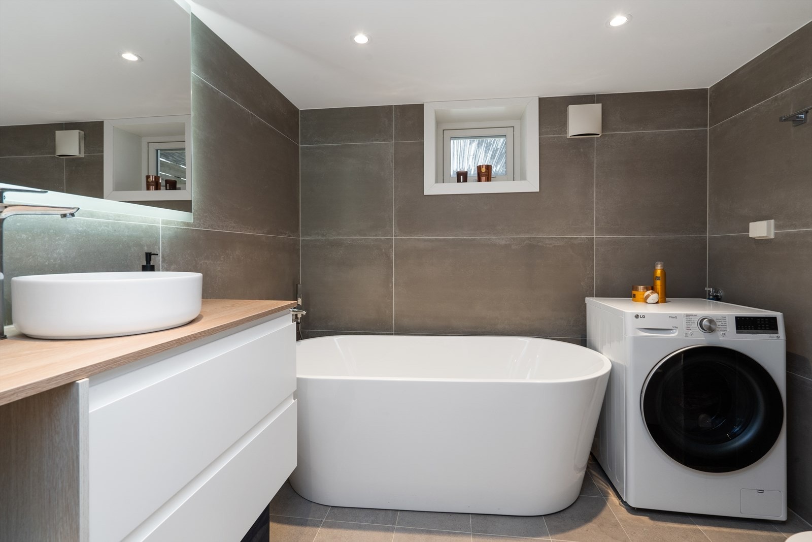 Kjellerbad med moderne innredning, romslig badekar og opplegg for vaskemaskin. Det er varmekabler i gulvet. Galleribilde