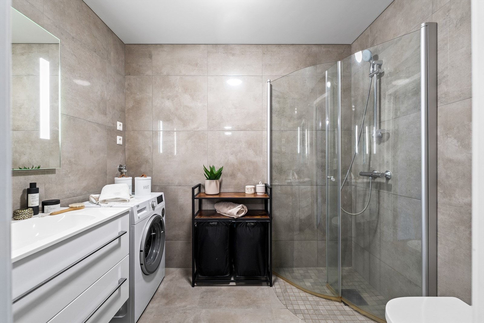 Flislagt baderom/vaskerom med varme i gulv. Baderommet er innredet med nedfelt servant, veggmontert toalett, dusjvegger/hjørne og opplegg for vaskemaskin. Galleribilde