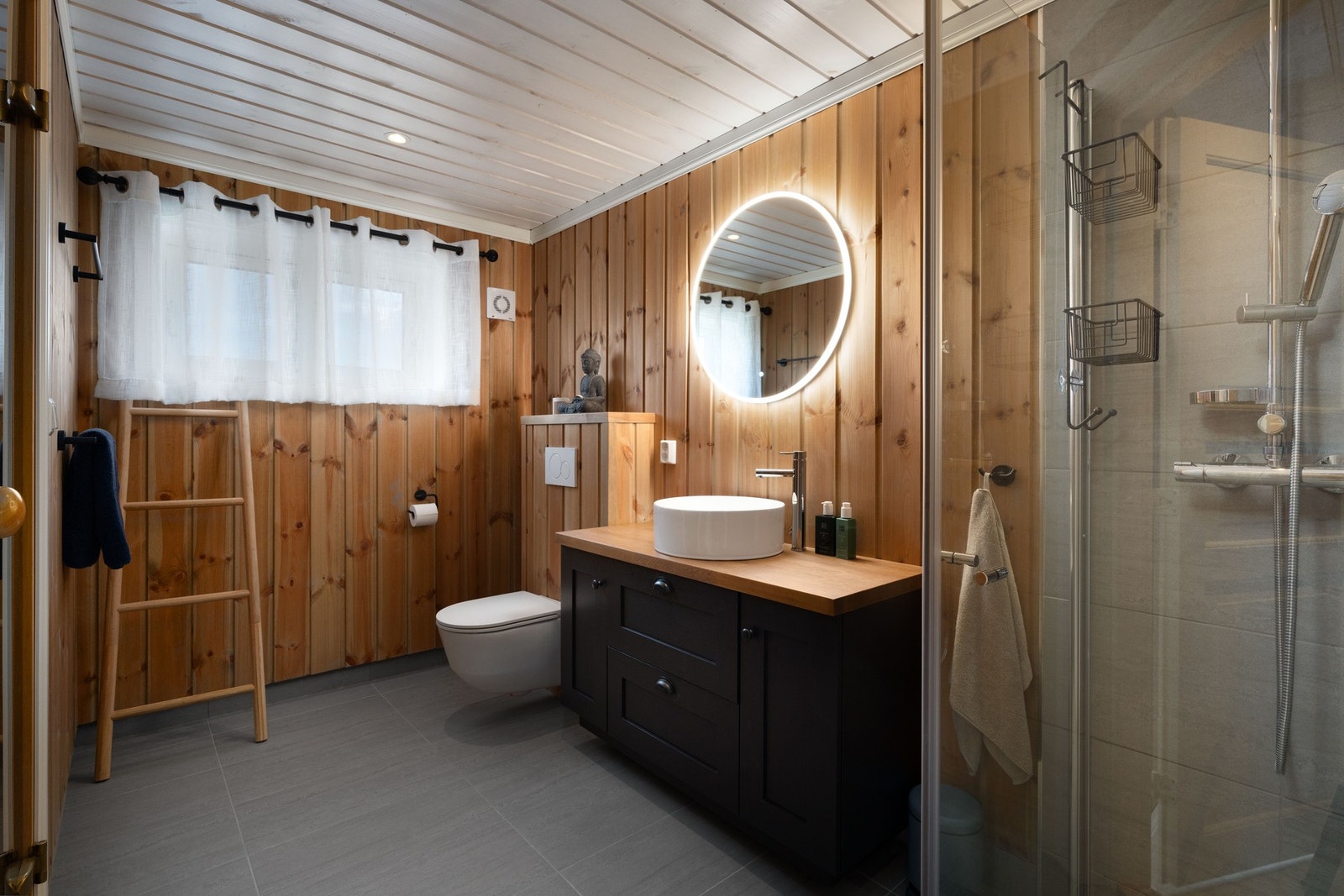 Innbydende bad med fliser på gulv og vegger i dusjhjørne. Det er montert innfellbare dusjdører, justerbar takdusj og hånddusj.
Veggmontert toalett med innebygget sisterne, servantinnredning i heltre med toppmontert vask. Rundt speil med integrert ledlys. Galleribilde