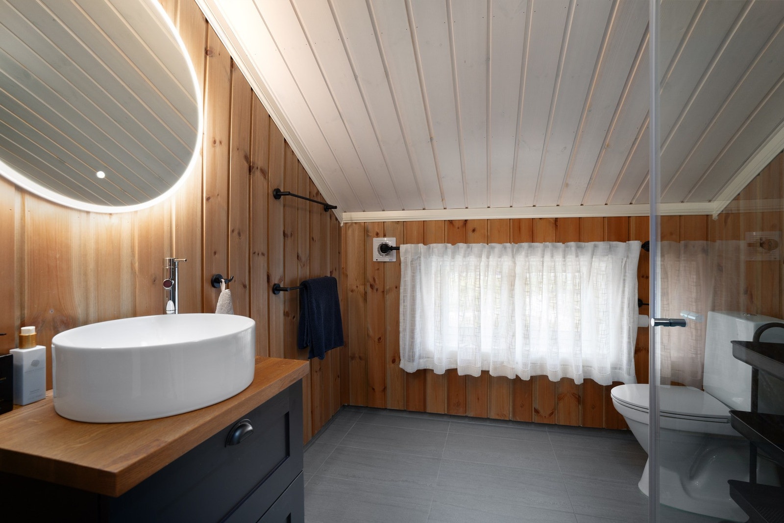 Badet i 2. etasje holder den samme stilen som hovedbadet.
Her er det fliser på gulv, innfellbare dusjdører, gulvmontert toalett, servantinnredning i heltre med toppmontert vask og rundt speil med ledlys. Galleribilde