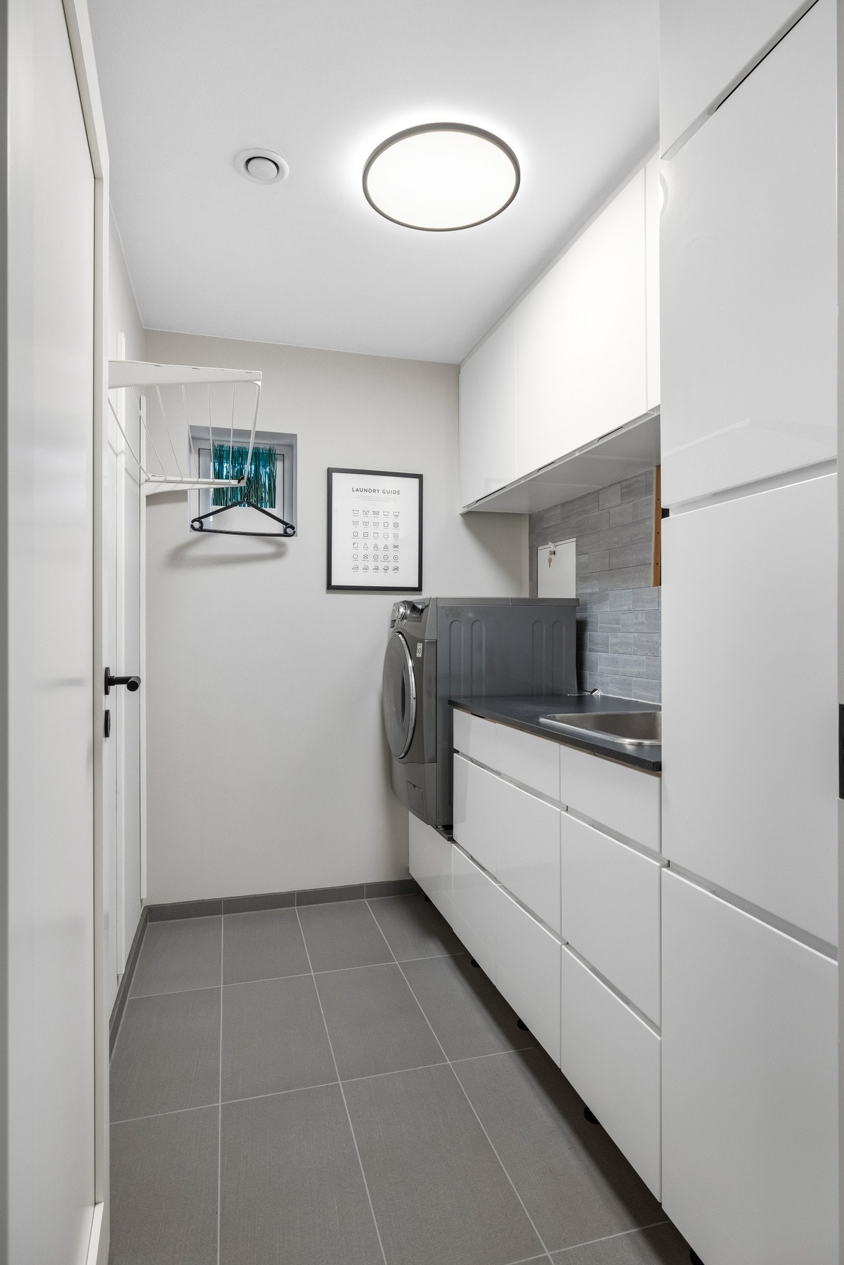 Vaskerommet har flislagt gulv med varme, og langs den ene veggen er et moderne benkeskap montert med nedfelt utslagsvask, matchende veggskap og tilhørende høyskap for praktisk oppbevaring. Galleribilde