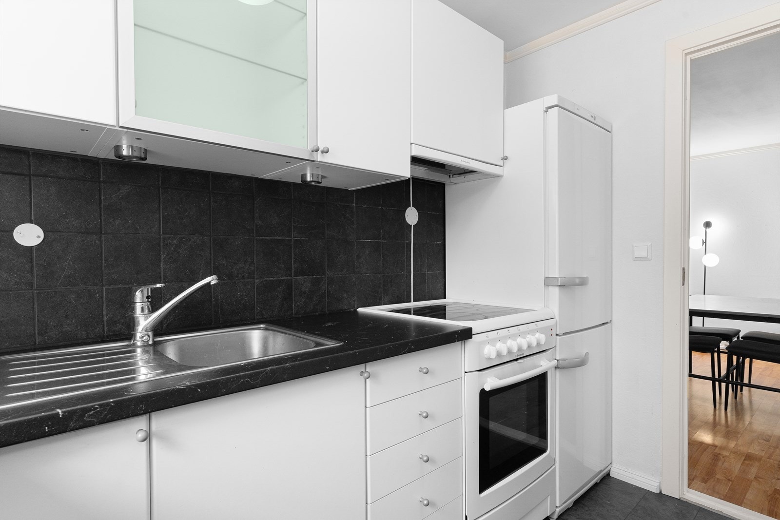 Stilren kjøkkeninnredning med hvite, glatte fronter og benkeplate av laminat. Kjøkkenet er utstyrt med hvitevarer som kjøleskap, oppvaskmaskin og komfyr. Det er kjøkkenventilator med avtrekk ut. Galleribilde