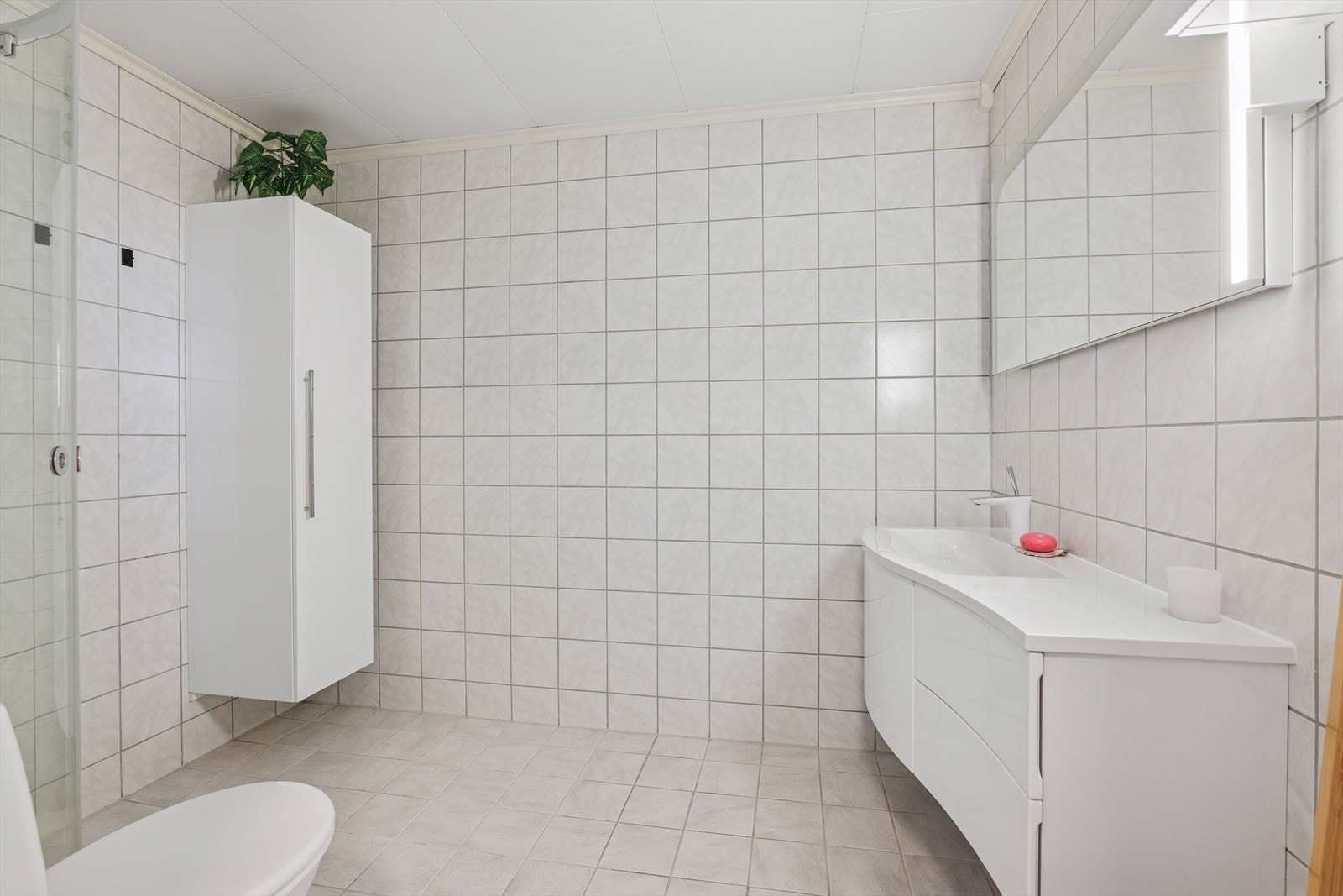 Badet i boligen er pent innredet med en stor baderomsinnredning med nedfelt servant og skuffeseksjon, toalett, belyst speil montert på vegg, veggmontert høyskap for fin oppbevaringsplass og et praktisk dusjkabinett for en enkel og sikker hverdag. Galleribilde