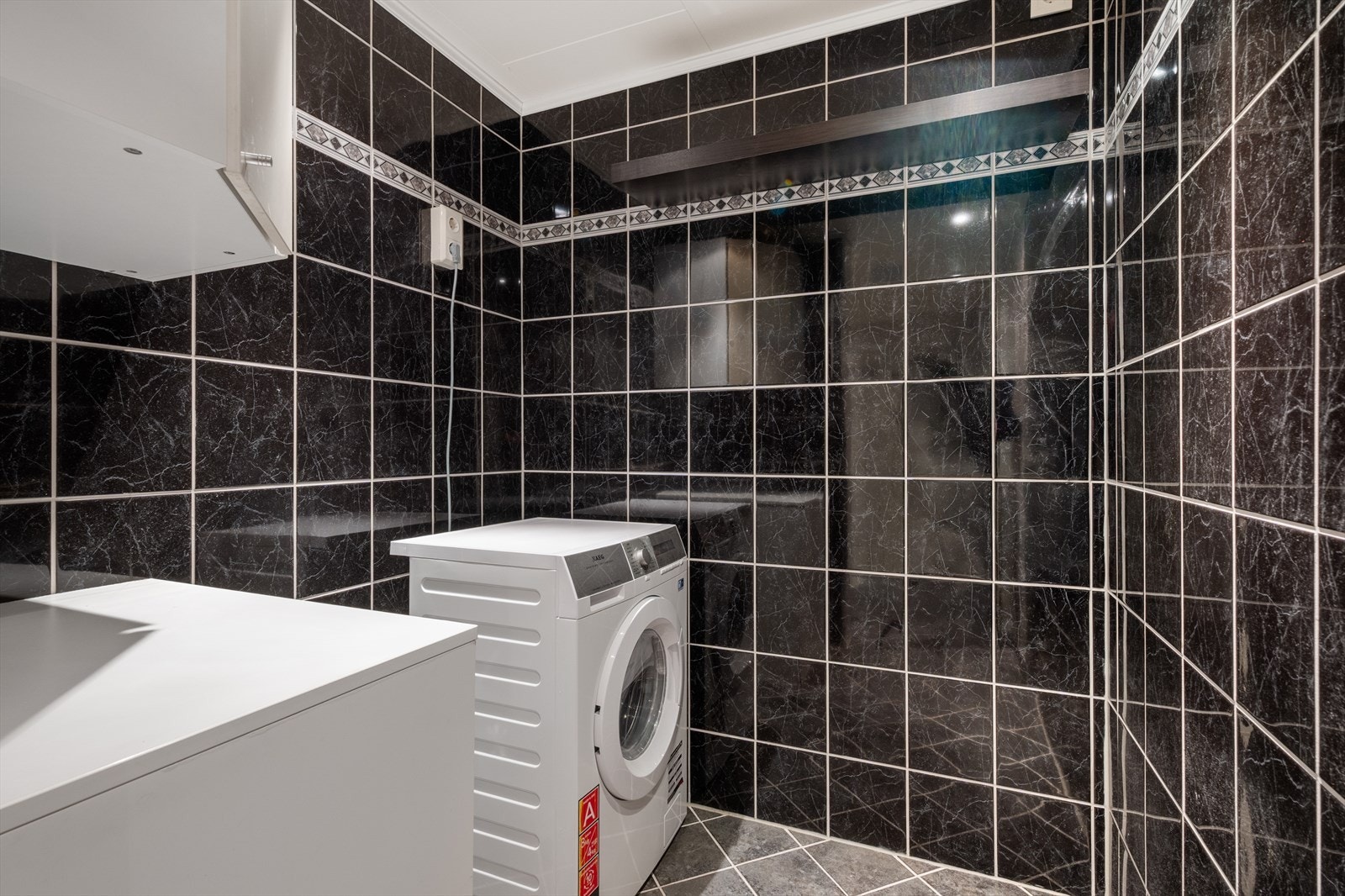 Vaskeromsdel med mørke fliser og opplegg for vaskemaskin. Galleribilde