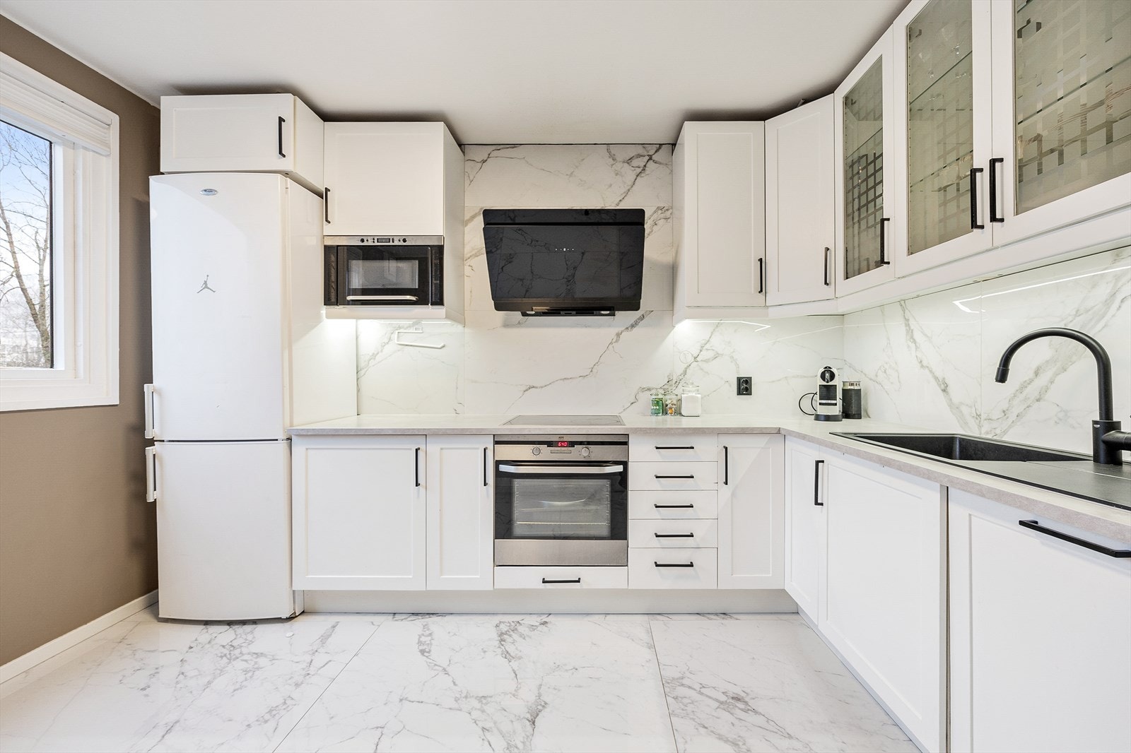 Moderne og stort kjøkken fra 2023 med hvite fronter og mørke håndtak. Benkeplaten er i laminat, og veggen over er kledd med plater i marmor-utseende. Galleribilde