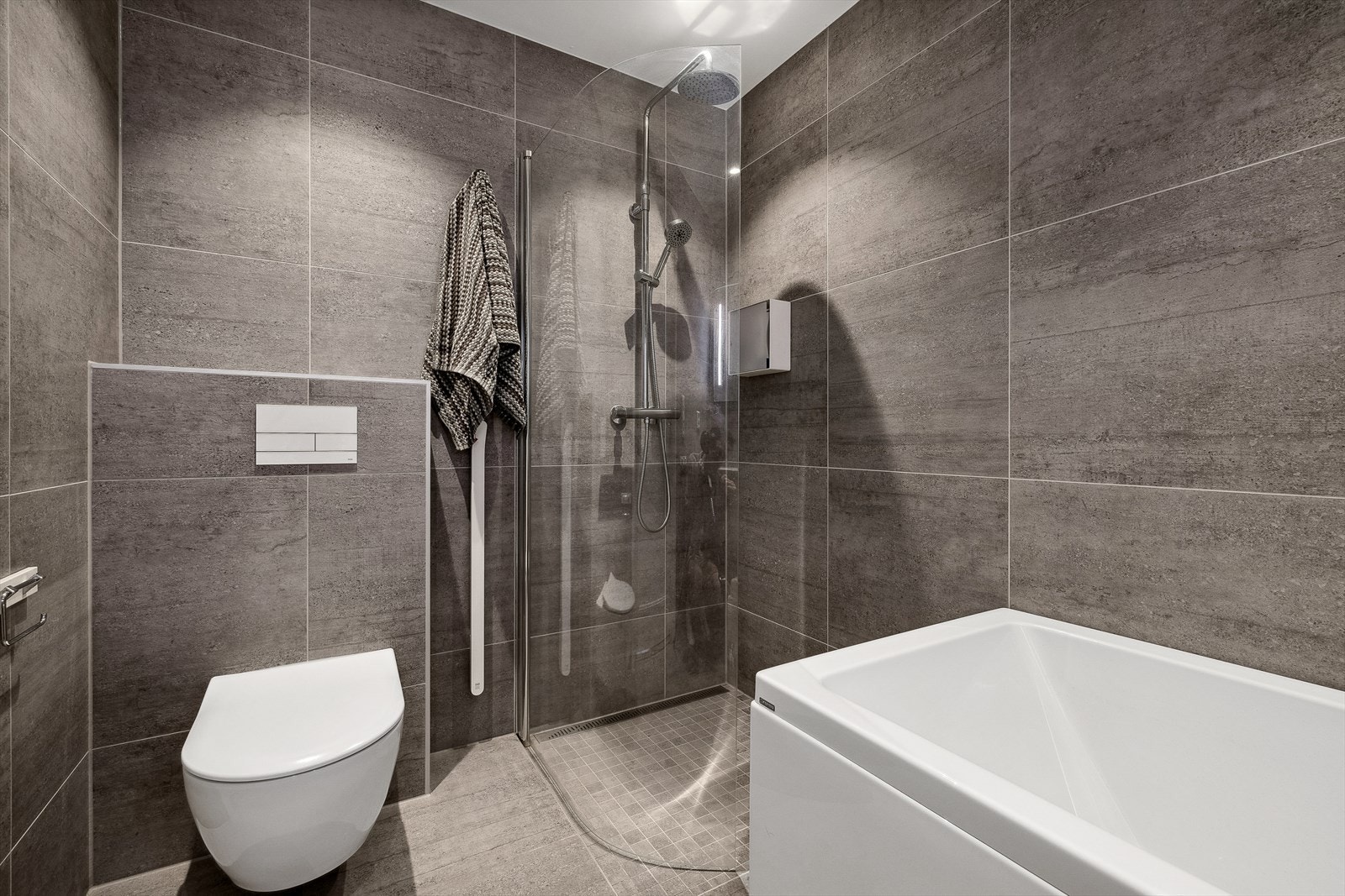 Bad i underetasjen er delikat med 60*60 fliser på vegger og gulv, varmekabler i gulv og downlights i himling. Badet er innredet med vask i seksjon, speil med belysning, badekar, vegghengt toalett og dusjhjørne. Galleribilde