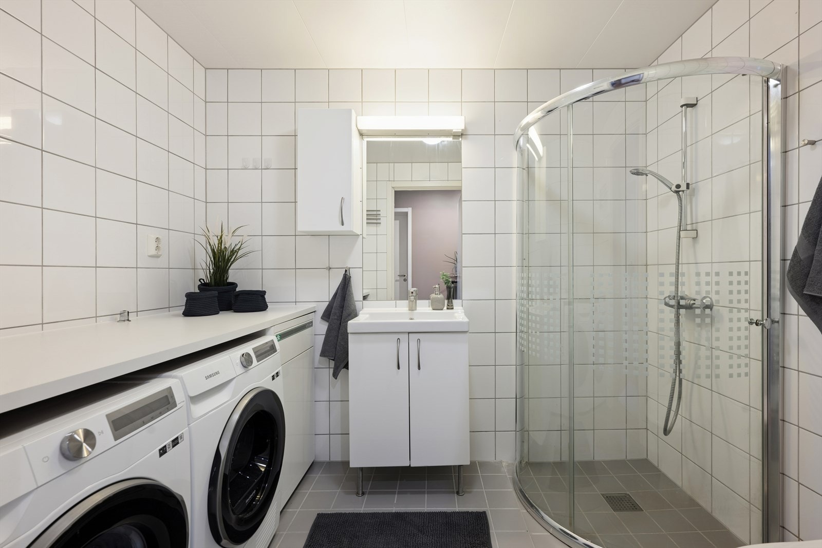 Badet er et stort og pent kombinert bad- og vaskerom, gjennomført med fliser på både gulv og vegger. I tillegg finnes en praktisk arbeidssone med benkeplate og opplegg for både vaskemaskin og tørketrommel. Galleribilde