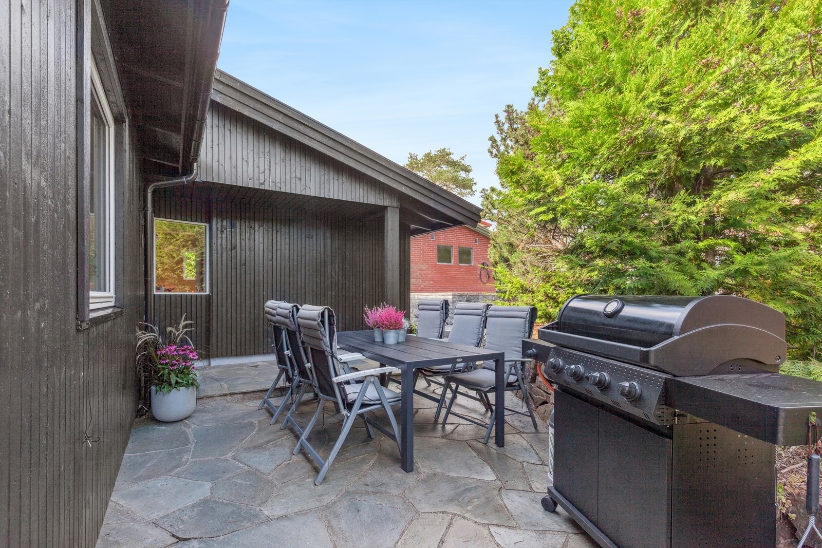 I tillegg gir et skiferbelagt uteområde ved inngangspartiet et eksklusivt og tidløst preg, ideelt for rolige kaffestunder. Kombinasjonen av tre og skifer skaper variasjon i uttrykk og funksjon, og gir eiendommen et gjennomtenkt og helhetlig utemiljø. Galleribilde