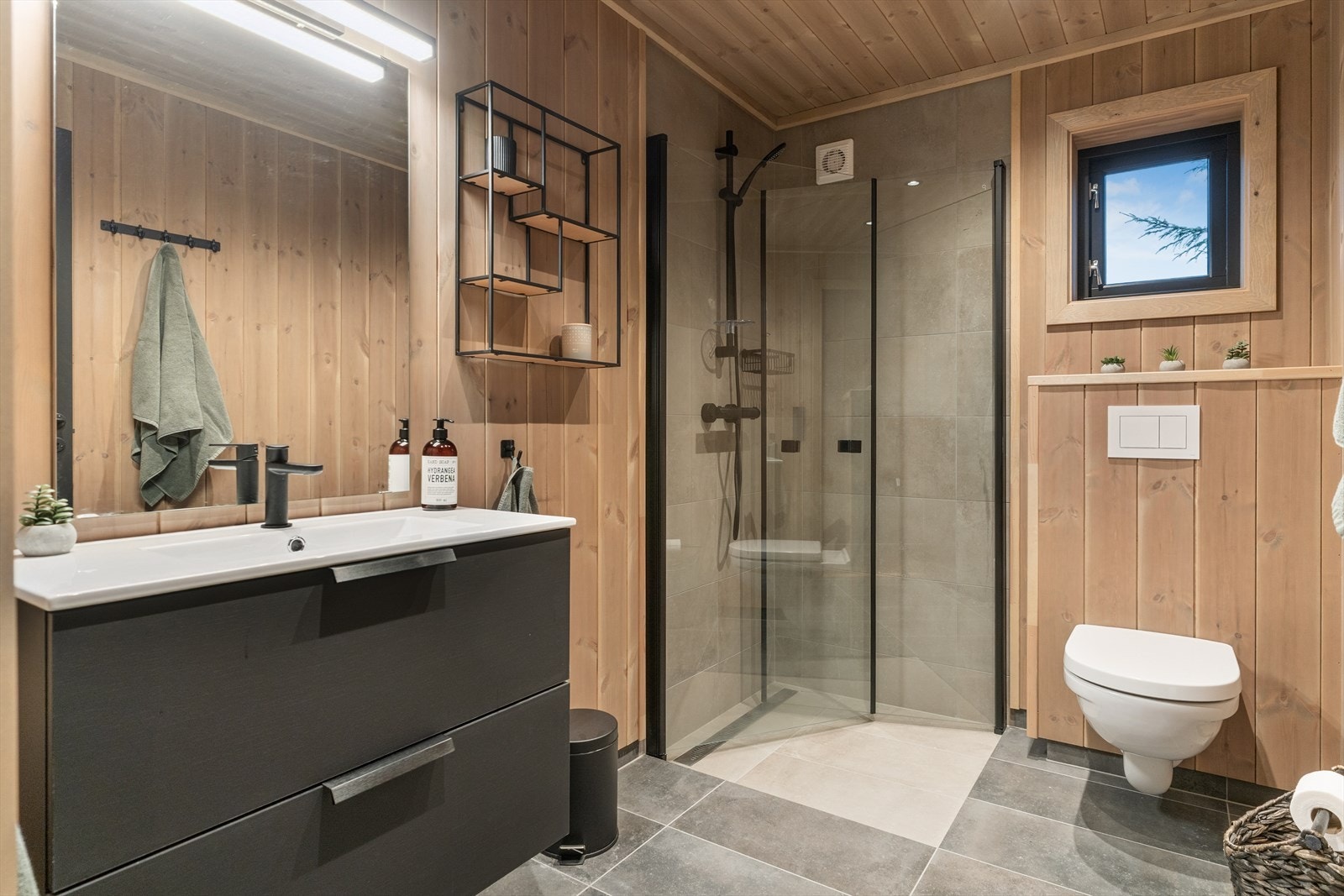 Baderom med flis på golv og i dusjsona. Det er panel på veggar og i himling. I dusjsona er det innfellbare dører, vegghengt toalett og skuffeinnreiing med heilstøypt servant. Her er det varmekablar. Galleribilde