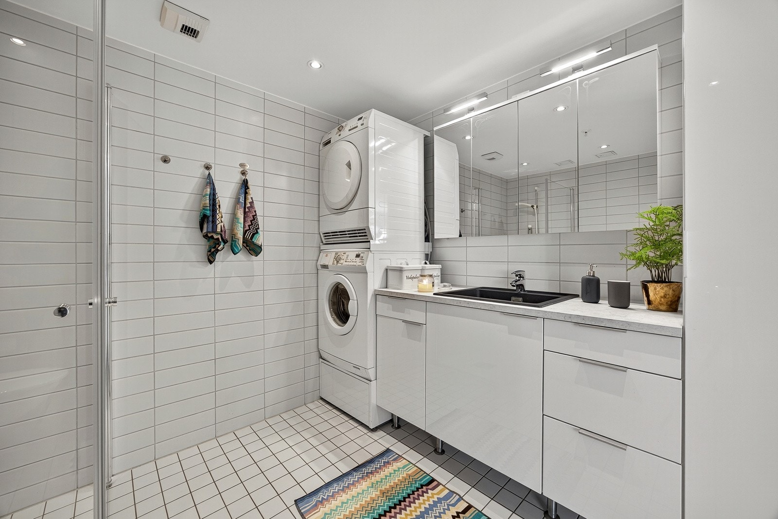 Baderomsinnredningen består av servantmøblement med glatte fronter og tilhørende veggskap, samt speilskap med belysning. Videre har badet dusjhjørne med innfellbare dører i herdet glass og veggfestet toalett. Opplegg til vaskemaskin og tørketrommel. Galleribilde