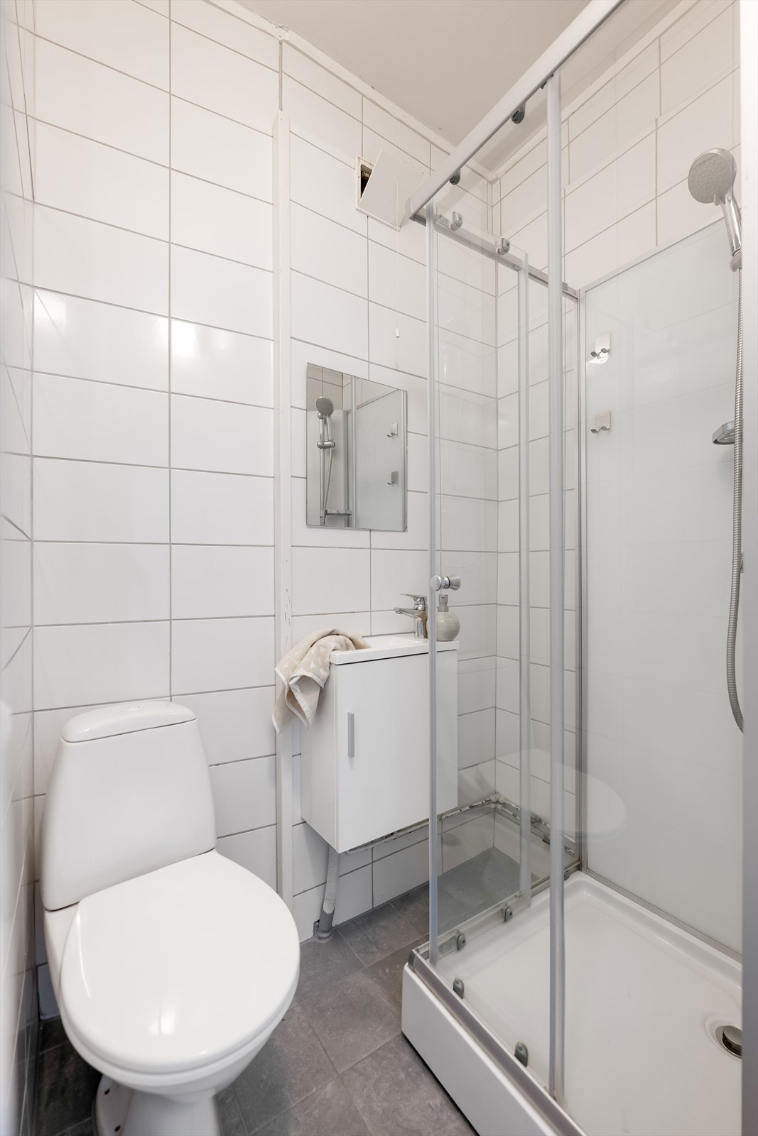 Flislagt baderom med servant m/ tilhørende underskap, speil, et gulvmontert klosett og et dusjkabinett. Galleribilde