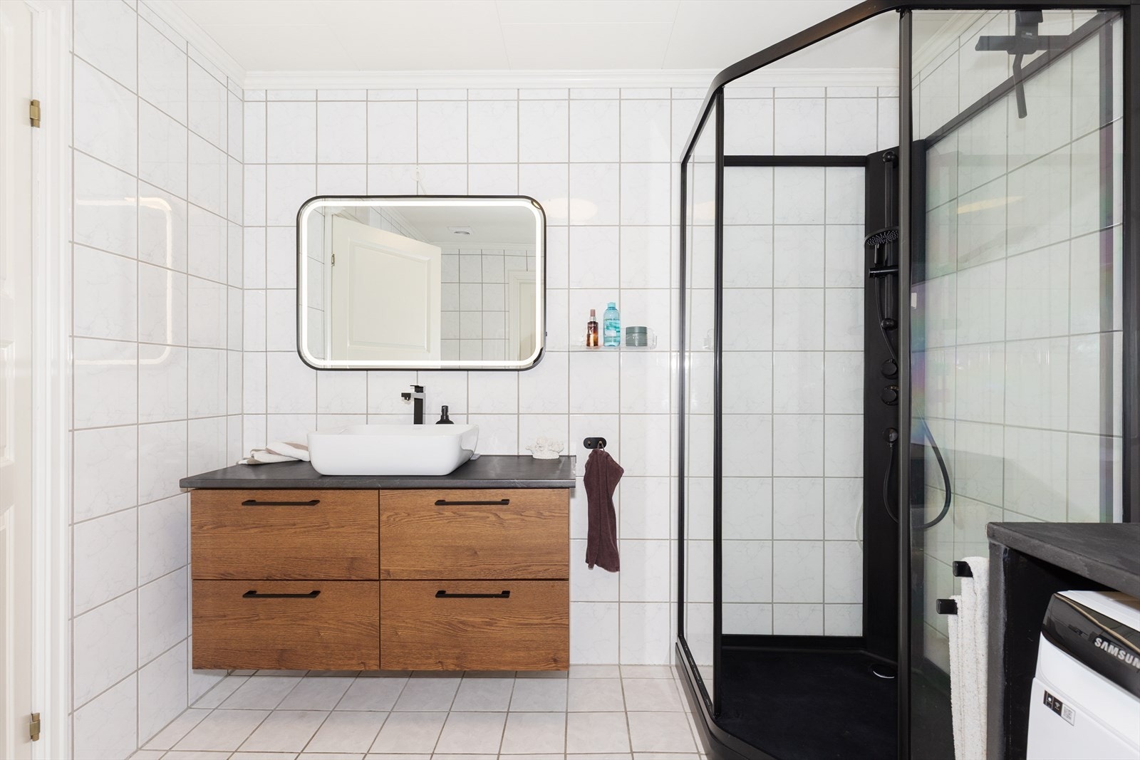 Badet fra byggeår er helfliset med varmekabler i gulvet. Sorte profiler og detaljer gir badet et eksklusivt helhetsinntrykk, og speilet på veggen gir et stilrent uttrykk kombinert med varm og behagelig belysning. Galleribilde