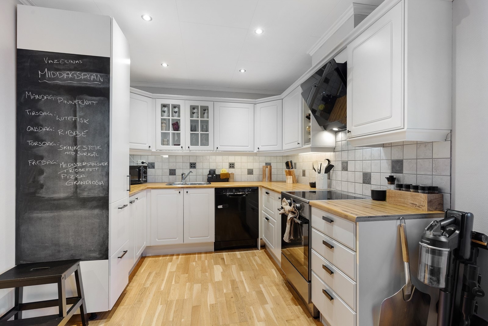 Kjøkkenet har avsatt plass til frittstående hvitevarer som oppvaskmaskin, komfyr og kjøl/fryseskap, og over kokesonen er det en stilig, skråstilt ventilator fra 2023 med avtrekk ut. Galleribilde