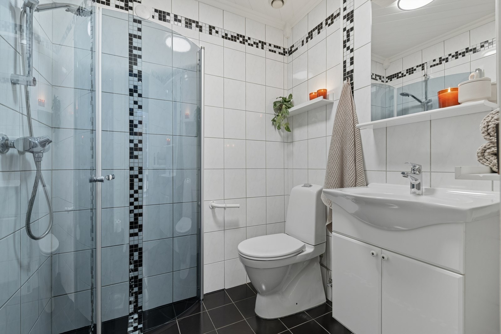 Baderommet i første etasje er innredet med mørke fliser på gulv og en kombinasjon av hvite og mørke fliser på vegg. I gulvet har man også varmekabler for ekstra komfort. Badet er ellers utstyrt med dusjhjørne, toalett og baderomsinnredning med servant. Galleribilde