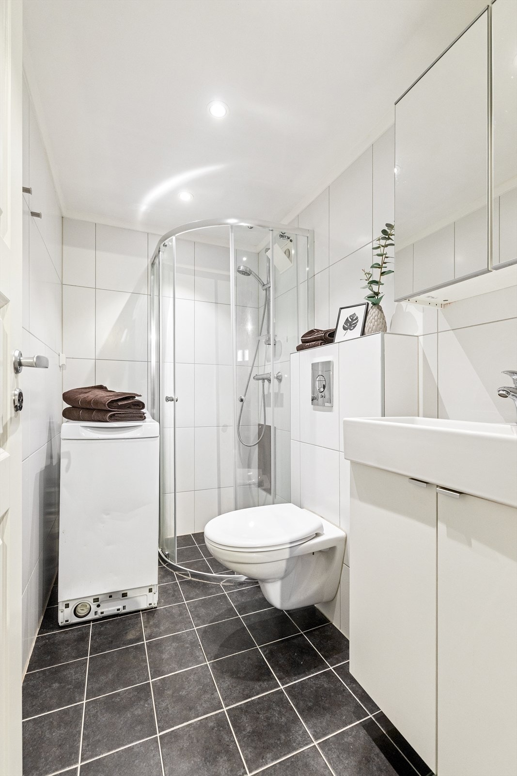 Hybeldel - Badet er utstyrt med dusjhjørne med innfellbare glassdører, veggmontert toalett og baderomsinnredning med skap, overlimt servant med ettgreps armatur og speilskap på vegg. Galleribilde