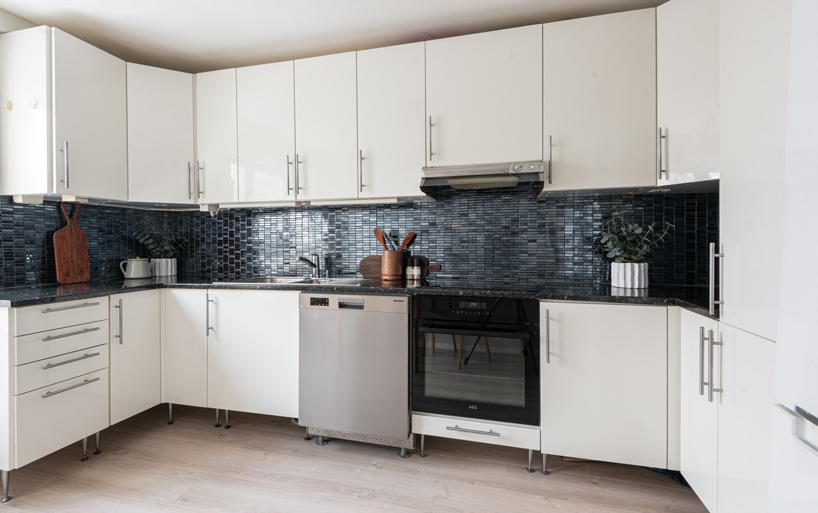 Kjøkkeninnredning med glatte fronter, fliser på veggen over benkeskapene og laminatbenkeplate med nedfelt kum i rustfritt stål. Kjøkkenet er utstyrt med ventilator Galleribilde