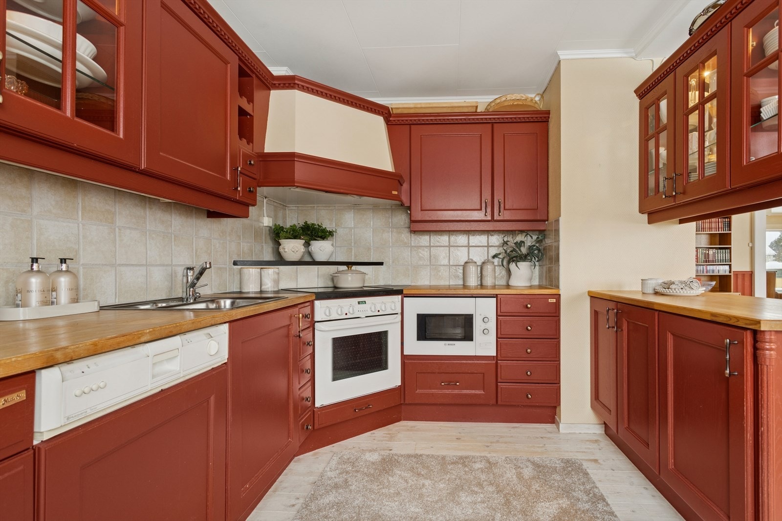 Fliser over arbeidsbenk og våtsone gir et rent og klassisk uttrykk. Overskap med glassdører tilfører et moderne preg, og den åpne kjøkkenøya skaper en naturlig overgang til stue og spisestue - perfekt for sosialt samvær. Galleribilde