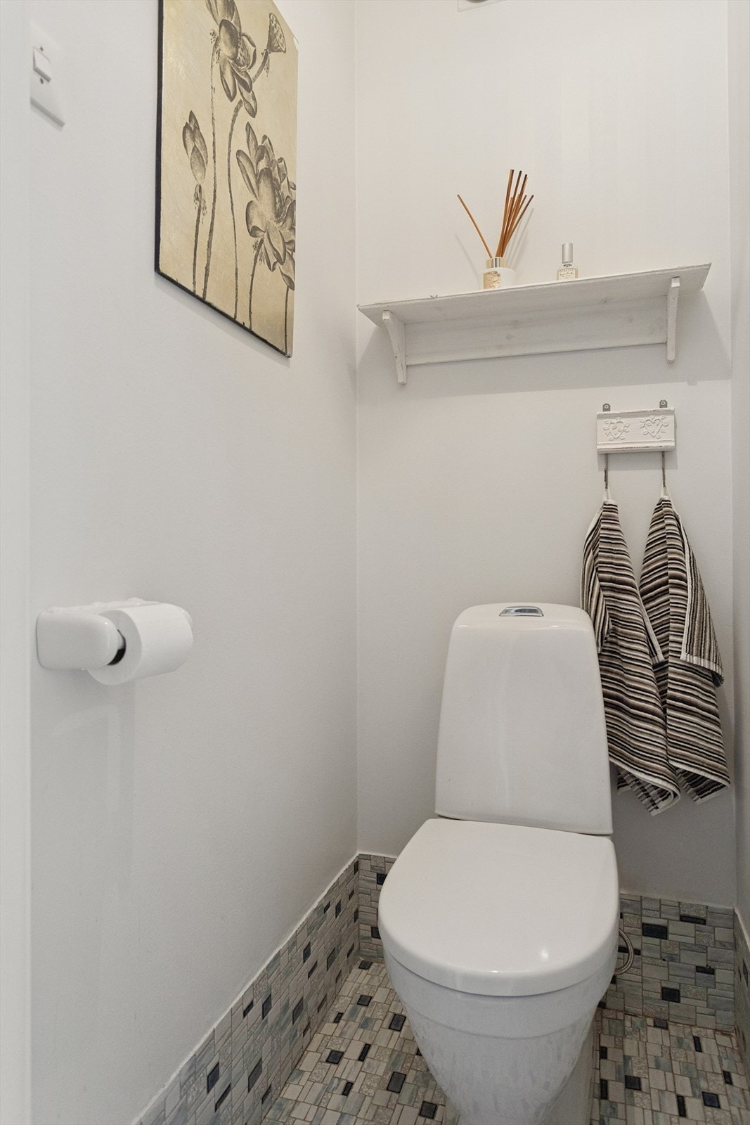 Toalettrom med mosaikkfliser på gulv. Hvite vegger gir et lyst uttrykk. Utstyrt med gulvmontert toalett og praktisk vegghylle. Galleribilde