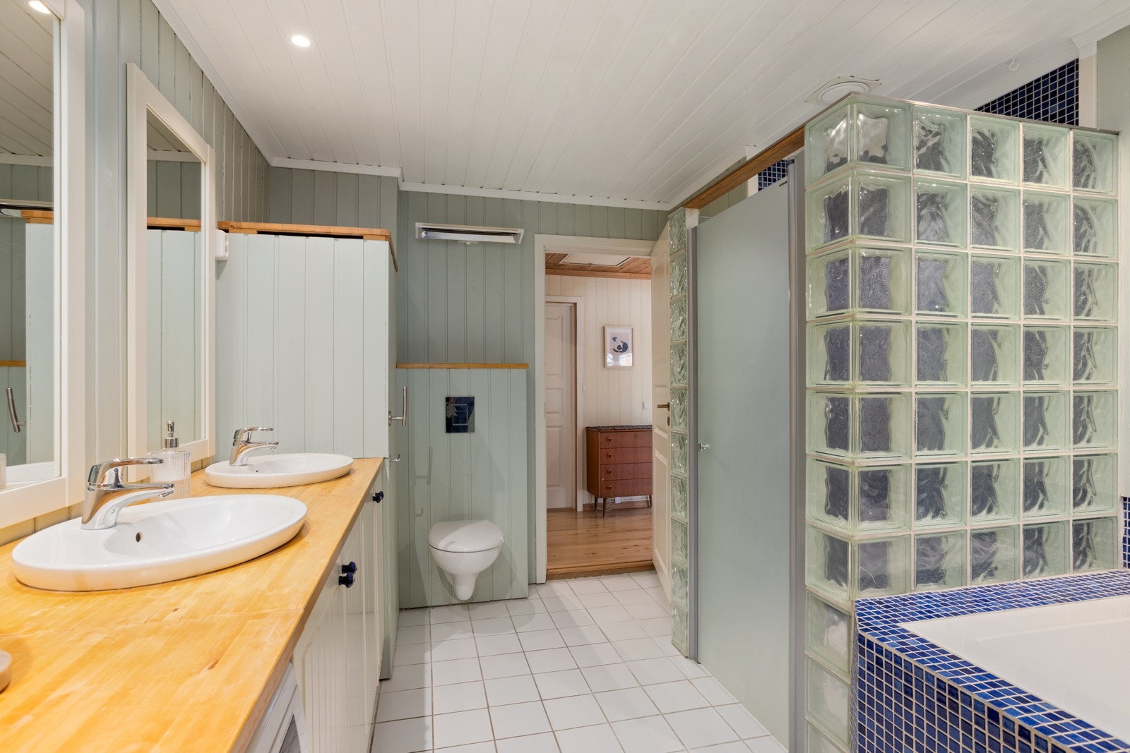 Baderomsinnredningen består videre av en romslig dusjnisje med glassvegg og glassdør med vegghengt dusjarmatur, vegghengt toalett og servantinnredning Galleribilde