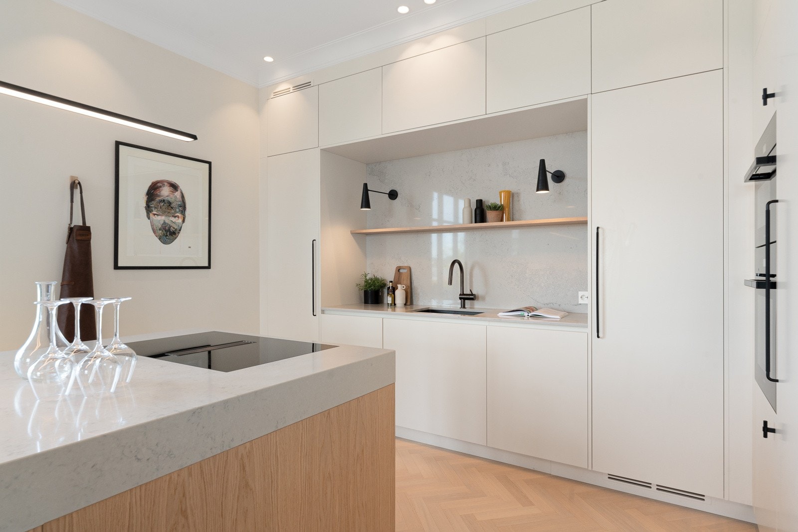 Innredningen langs veggen har glatte, lyse fronter, sorte håndtak og en inntrukket benk med benkeplate i lys, marmorert Caesarstone.