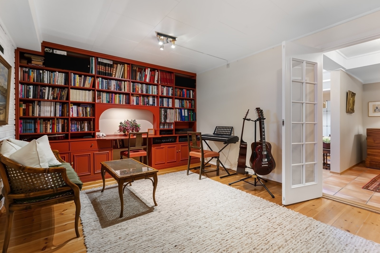 Plassbygde, rødmalte bokhyller fra gulv til tak langs to vegger, med integrert arbeidspult og skap. Galleribilde