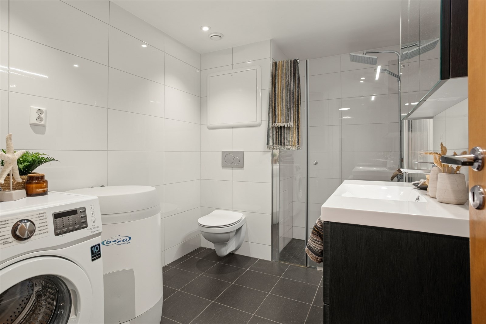 Rommet er utstyrt med en servant med underskap, et speilskap med belysning, et vegghengt toalett og en dusjsone med glassdør. Det er også plass og opplegg for vaskemaskin. Galleribilde