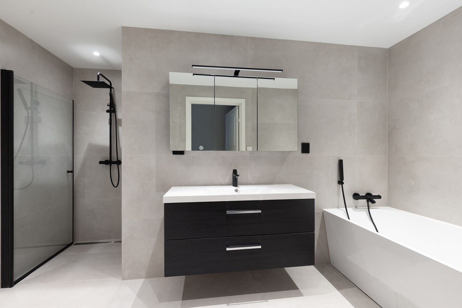 Lekkert bad med flis på gulv og vegger, og sensorstyrte spotter i tak. Her er det vegghengt toalett med lekkasjepåviser, badekar, dusjhjørne med vegghengt armatur og dør i glass, samt innredning. Galleribilde