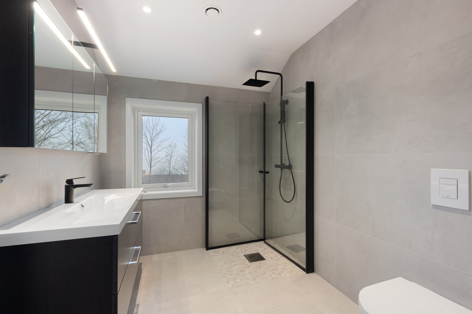 Flott bad med flis på gulv og vegger. Vegghengt toalett med lekkasjepåviser, dusjhjørne med armatur på vegg og dører i glass, samt innredning med heldekkende hvit vask, underskuffer med slette fronter og overskap med speildører og lys. Varmekabler. Galleribilde