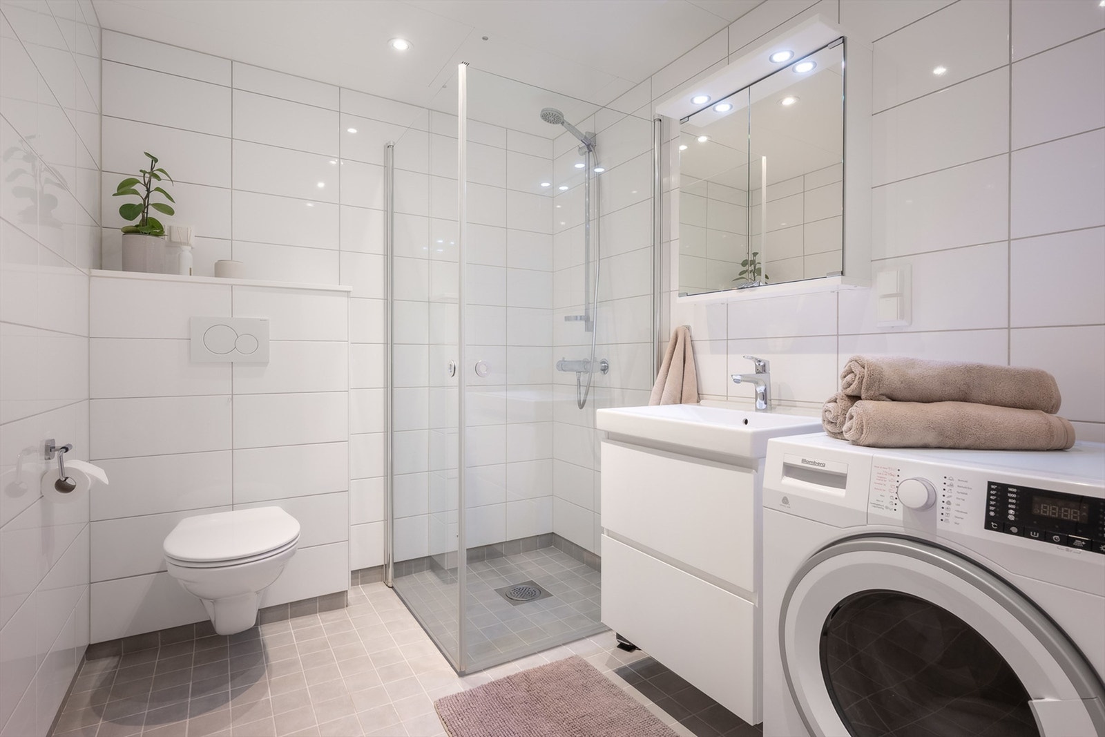 Badet er pent flislagt på gulv og vegger med vannbåren gulvvarme. Badet er innredet med dusj på gulv med dusjdører i herdet glass, servant med skuffeinnredning og vegghengt klosett. Det er både plass og opplegg for vaskemaskin og tørketrommel. Galleribilde